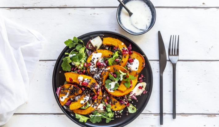 Fotografia vista de cima de uma salada de folhas com molho de laranja, pedaços de abóbora, sementes e um ramo de coentro em um prato raso preto. Ao lado tem talheres e um molho de iogurte em um pote pequeno de vidro azul, com uma colher apoiada.