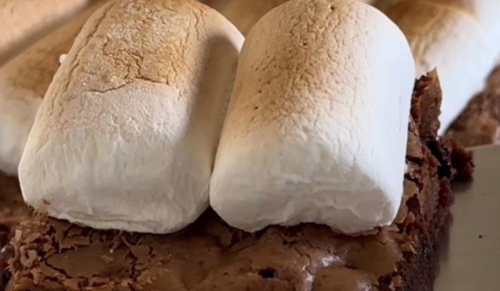 Fotografia de pedaços de brownie com marshmallow assados por cima sobre uma mesa branca.