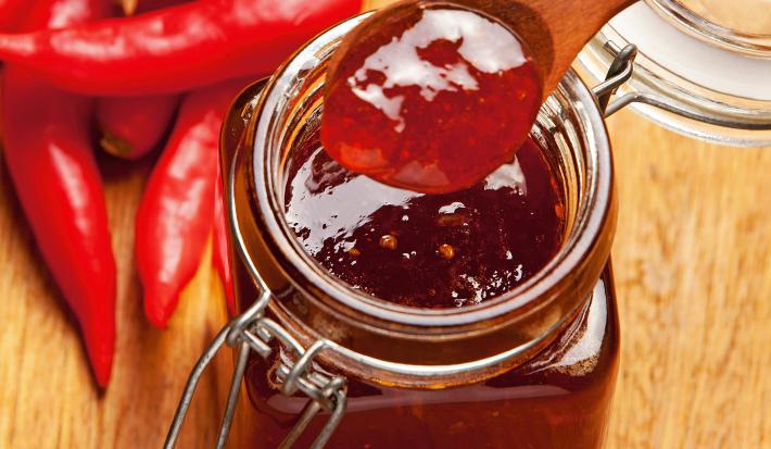 Fotografia de um recipiente de vidro decorativo com geleia de pimenta e laranja. O recipiente está sobre uma mesa de madeira, ao lado de várias pimentas dedo-de-moça.