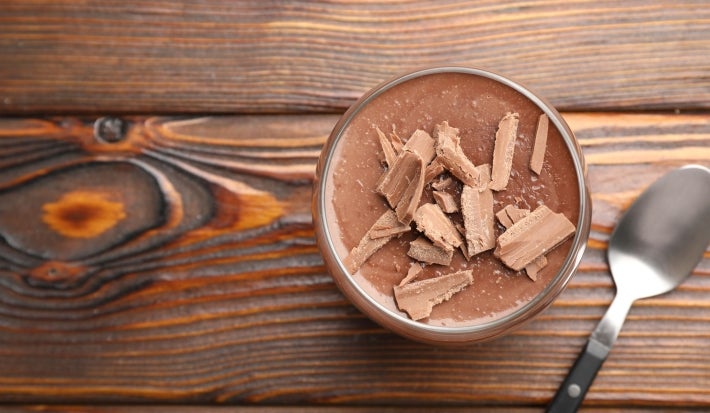 Fotografia em tons claros, vista de cima, de um recipiente de vidro pequeno com a sobremesa, finalizada com ganache e raspas de chocolate sobre uma mesa de madeira. Ao lado tem uma colher de sopa.