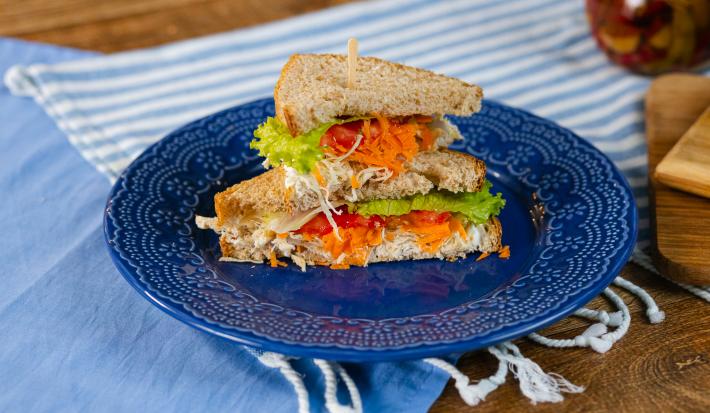Fotografia em tons de azul com um prato azul ao centro. Em cima do prato existe uma receita de sanduíche natural recheado com frango desfiado, creme de ricota, cenoura, alface e tomate