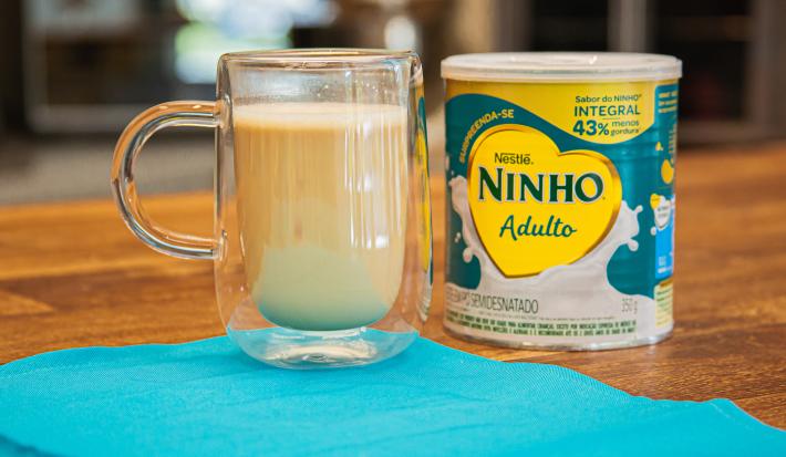 Fotografia em tons de azul com uma caneca de virdro ao centro. Dentro da caneca existe um cappuccino de cor caramelo e ao lado existe uma lata de leite NINHO Adulto em pó