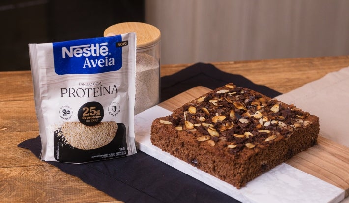 receita de bolo proteico molhadinho de banana e chocolate servida em uma porção grande sobre uma mesa de madeira decorada com um pote e uma embalagem de aveia proteína