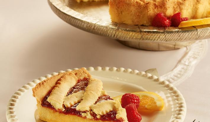 Fotografia em tons de bege em uma mesa de madeira, um guardanapo de pano bege e branco, um prato redondo branco com detalhes bege e uma fatia da crostata com recheio de laranja. Ao fundo, um prato redondo maior com a crostata inteira.