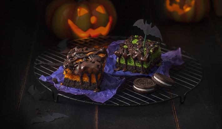 Fotografia em tons de roxo e laranja em uma bancada preta com desenhos de morcegos e abóboras desenhadas ao fundo. Ao centro, um suporte de grelha preto com os pedaços de bolos de chocolate com Negresco em cima.