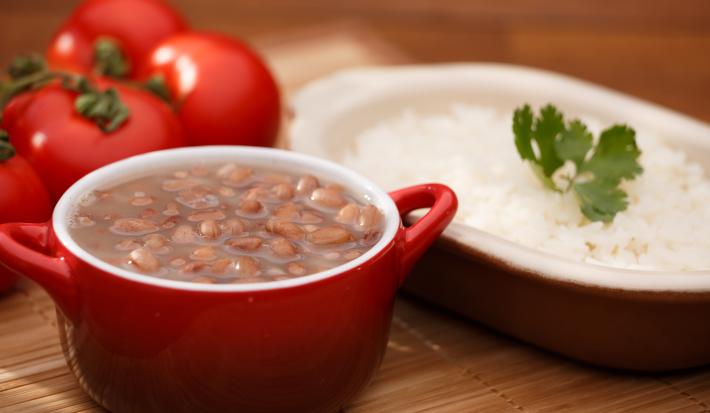 foto em tons de marrom, vermelho e branco de uma bancada marrom vista de frente. Contém um recipiente oval com arroz e outro redondo vermelho com alças que contém feijão.