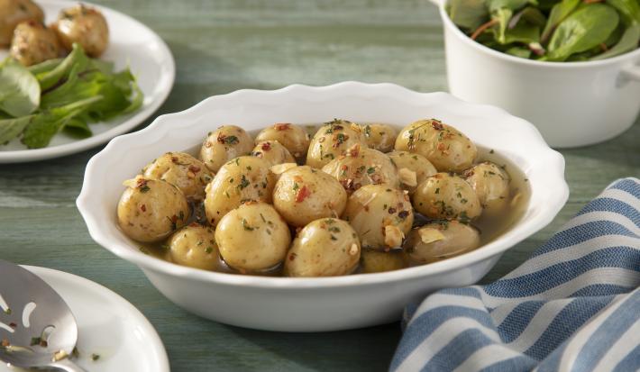 Fotografia em tons de azul em uma bancada de madeira verde, um pano branco com listras azuis, um recipiente oval branco com várias batatas bolinhas com pimenta calabresa.