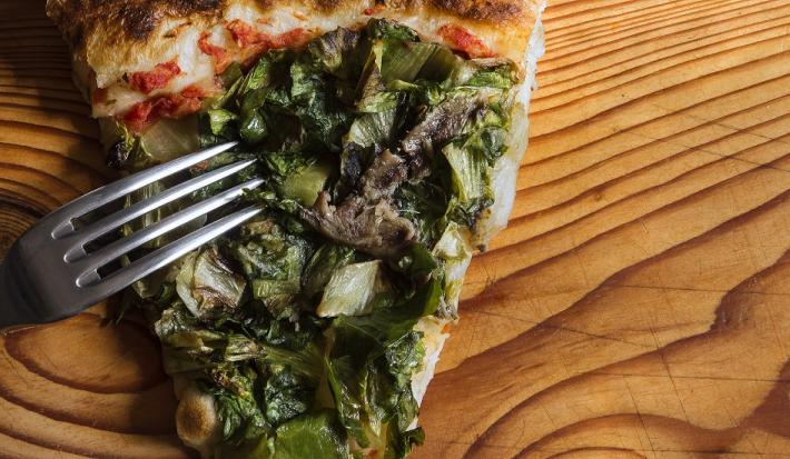 Fotografia em tons de verde em uma bancada de madeira de cor marrom. Ao centro, uma fatia de pizza de escarola com um garfo ao lado.