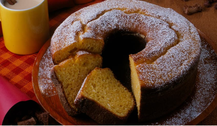 Fotografia em tons de vermelho em uma bancada de madeira escura com um pano laranja e vermelho xadrez, um suporte para bolo com o bolo de milho em cima dele cortado em algumas fatias. Ao lado, xícaras coloridas com cappuccino dentro.