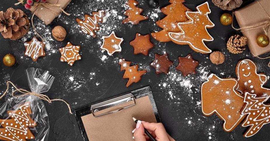 Mesa com biscoitos de Natal decorados, presentes embalados, e uma mão escrevendo em um papel.
