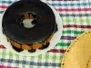 Um bolo de cenoura inteiro com cobertura de chocolate, no formato redondo com furo no meio, está em um prato branco sobre uma toalha colorida xadrez. Um chapéu de palha aparece parcialmente no canto da imagem.