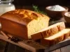 Fotografia de um bolo recém-assado em uma tábua de madeira, com uma crosta dourada e interior macio. O cenário rústico inclui tigelas com farinha e açúcar, além de especiarias.
