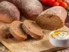 Fotografia de uma tábua média de madeira sobre uma mesa de madeira escura. Sobre a tábua tem uma receita de pão australiano com três fatias cortadas e sobrepostas ao lado de dois pedaços maiores do pão.