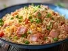 Em cima de uma mesa, prato redondo com porção de arroz com presunto cortado, ervilha, milho, decorado com cebolinha picada.