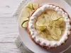 Fotografia em tons mais claros de uma torta inteira de limão com raspas e rodelas finas de limão. O chantilly está apenas na borda da torta, ao lado dela tem três fatias, que está sobre um prato grande e raso de vidro.