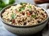 Em cima de uma mesa, tigela com arroz soltinho uvas-passas pretas e brancas, decorado com folhas de cheiro-verde.