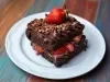 Sobre uma mesa, prato com uma fatia de bolo de chocolate. A massa é de chocolate, recheada com chocolates e morangos picados, coberta com chocolate e decorada com granulado e morangos cortados ao meio.