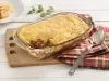 Fotografia em tons de amarelo e branco de uma bancada de madeira branca, tábua de madeira, pano quadriculado vermelho e marrom e uma travessa com o escondidinho, uma fatia retirada, mostrando a carne desfiada e a camada de purê. Ao lado colher prateada.