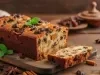 Fotografia de um bolo de frutas recém-assado, com crosta dourada e rica em passas e frutas secas. A fatia cortada revela a textura úmida, em um ambiente rústico com elementos decorativos como anis estrelado e canela.