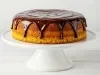 Um bolo de cenoura inteiro, com cobertura de chocolate brilhante escorrendo pelas laterais, sobre um prato de pedestal branco. O bolo tem uma aparência alta, dourada e bem assada, com cobertura uniforme no topo.