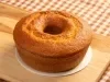 O bolo de cenoura simples com furo no meio e aparência bem fofinha. Ele está em uma base branca sobre uma tábua de madeira, com uma toalha de mesa xadrez ao fundo, criando um clima caseiro e acolhedor.