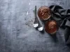 Fotografia vista de cima em tons escuros de dois copos de vidro com a sobremesa, finalizada com a ganache e raspas de chocolate. Ao redor tem duas colheres de sobremesa e um pano dobrado preto, sobre uma mesa de tom de cimento queimado escuro.
