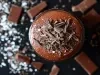 Fotografia em tons mais escuros, vista de cima, de um pote de vidro com a sobremesa, finalizada com ganache e raspas de chocolate. O pote está sobre uma mesa preta com pedaços de chocolate.