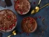 Fotografia em tons escuros, vista de cima, de três recipientes de vidro, grandes, sobre uma mesa de tom escuro, com algumas colheres douradas, raspas e pedaços de chocolate. Dentro dos recipientes tem a sobremesa, finalizada com ganache e raspas.
