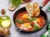 Fotografia em tons claros de uma receita de Shakshuka com pães por cima dentro de uma frigideira de ferro preta sobre uma mesa cinza. Ao lado da frigideira tem um recipiente com azeite, uma mini tábua de madeira e um moedor de pimenta de madeira.