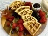 Foto em tons de bege e marrom da receita de waffle delícia servida em seis porções sobre uma tábua de madeira, com frutas vermelhas espalhadas pela tábua, além de um recipiente com geleia de frutas e vidro de mel