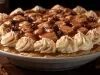 Em cima de uma mesa, refratário redondo com massa crocante, doce de leite, decorado com chantilly, bananas e uvas-passas.
