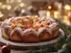 Fotografia de um bolo natalino circular, coberto com açúcar de confeiteiro e decorado com frutas secas coloridas. O fundo apresenta luzes suaves e elementos festivos.
