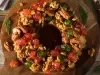 Fotografia de um bolo de frutas circular em forma de guirlanda, adornado com frutas cristalizadas e nozes, colocado sobre uma tábua de madeira rústica, cercado por ramos de pinheiro, evocando um clima natalino e aconchegante.