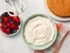 Fotografia em tons claros vista de cima de um recipiente claro fundo com creme de ninho apoiado em um pano colorido dobrado ao lado de um bolo inteiro de massa branca e um recipiente pequeno fundo com frutas vermelhas, sobre uma mesa branca.