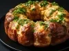 Fotografia de um bolo em formato de anel, com crosta dourada e brilhante, coberto com queijo derretido e ervas frescas. O bolo é apresentado em um prato preto.