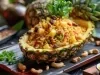 Em um abacaxi cortado ao meio, porção de arroz com abacaxi picados, ervilhas, uvas-passas e castanha-do-Pará.