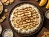 Em cima de uma mesa, refratário redondo com massa crocante, decorado com chantilly, bananas em rodelas e linhas de doce de leite.
