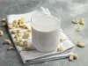 Fotografia em tons claros de um copo de vidro com uma bebida vegetal de castanha de caju apoiado em um pano branco dobrado com algumas listras escuras, que está sobre uma mesa de cimento queimado. Por cima da mesa e do pano tem castanhas espalhadas.