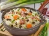Em um recipiente redondo cinza de porcelana, há uma base de arroz soltinho, combinado com pedaços de presunto cozido, coloridos vegetais, como cenoura, uvas-passas e decorado com cebolinha picada.