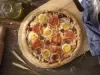 Fotografia em tons de azul em uma bancada de madeira escura, um paninho azul, uma tábua de madeira com a pizza portuguesa em cima dela. Ao lado, um potinho de azeite e um rolo de massa.