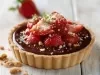 Fotografia de uma mini torta de bombom de morango, por cima tem ganahce de chocolate, alguns morangos decorando e um pouco da base triturada. A torta está sobre uma mesa de madeira clara, e ao fundo tem um morango inteiro.