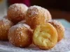 Fotografia de um prato com bolinhas de massa frita, cobertas de açúcar, com uma aparência dourada e crocante. Uma delas está cortada, mostrando um interior macio e fofo, dispostas sobre um delicado guardanapo de renda.