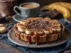 Em cima de uma mesa, prato com banoffee com massa de biscoitos crocante, bananas em rodelas, polvilhado com chocolate em pó.