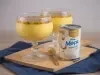 Foto da receita de Mousse de maracujá feita com o suco concentrado da fruta, servida em taças de vidro, com uma decoração de calda de maracujá com sementes, sobre uma tábua de madeira e ao lado de uma lata de Leite Moça