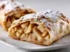 Fotografia em tons claros de um strudel de maçã cortado ao meio, sobre um prato branco raso. O strudel mostra o rechieo de maçã em pedaços com canela, e por cima tem açúcar de confeiteiro.