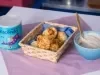 Fotografia em tons claros de uma cestinha de madeira retangular sobre um pano quadrado roxo. Dentro da cestinha tem um paninho menor azul com os pães de queijo, ao lado tem uma lata de Ascenda e um recipiente azul fundo pequeno com o creme de inhame.