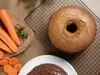 Bolo de cenoura com furo no centro, assado e dourado, resfriando sobre uma grade. Ao lado, cenouras frescas e um bolo decorado com cobertura de chocolate e granulados.