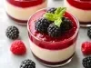 Fotografia de uma elegante sobremesa em um copo transparente, com uma base cremosa branca, coberta por um vibrante compota de frutas vermelhas, decorada com amoras frescas e uma folha de hortelã.