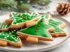 Em cima de uma mesa, prato redondo com biscoitos em formato de árvore de Natal, decorados com glacê verde e confeitos de bolinhas vermelhas.