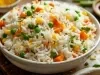 Em cima de uma mesa, tigela com porção de arroz branco soltinho com milho, cenouras e milho-verde, decorado com fatias de bacon e folha de salsa.
